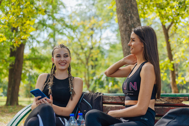 Chaud attrayant femmes souriant et glisser sur leur téléphone tandis que assis dans un parc et de prendre une pause de leurs activités sportives. - Photo, image