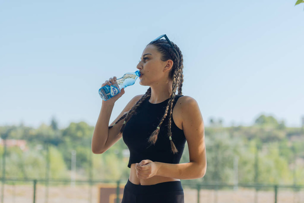 Retrato de una atleta feliz bebiendo agua de una botella de plástico después de un entrenamiento al aire libre - Foto, imagen
