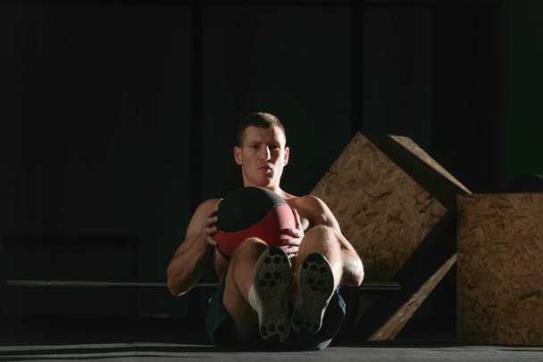 Lähikuva valokuva tumma kontrasti komea kehonrakentaja treenata istuen käyttäen lääkepallo - Valokuva, kuva