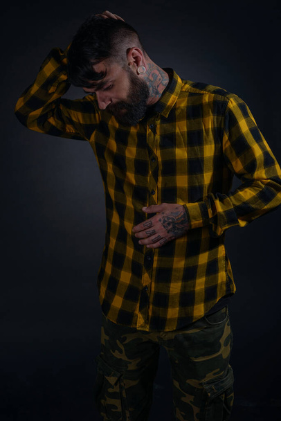 Привабливий чоловік з татуюваннями позує, одягнений у жовту та чорну картату сорочку на чорному тлі
 - Фото, зображення