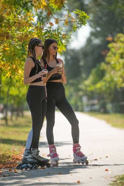 Two attractive best friends talking and drinking smoothies in the park while standing on their rollerskates - Photo, Image
