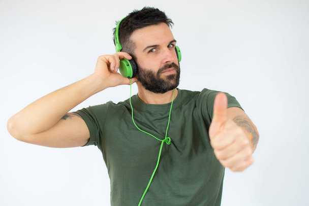 Young bearded man in headphones on head listening music, showing thumb up and standing isolated on white background with copy space - Zdjęcie, obraz