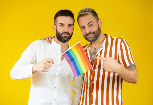 Feliz pareja gay sosteniendo la bandera homosexual sobre fondo amarillo. - Foto, imagen