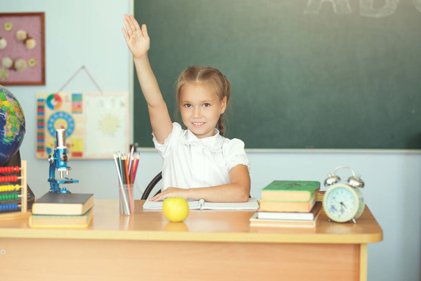 Cute schoolgirl pupil raising hand in classroom. - Фото, изображение
