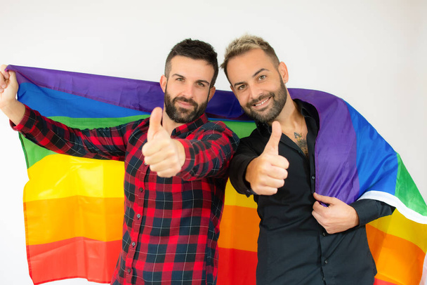 heureux gay couple enveloppé avec l 'homosexuel drapeau avec pouce jusqu' sur blanc fond. - Photo, image