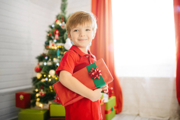 Netter kleiner Junge von etwa fünf Jahren mit einem Geschenk in einem geschmückten Weihnachtszimmer mit Weihnachtsbaum. - Foto, Bild