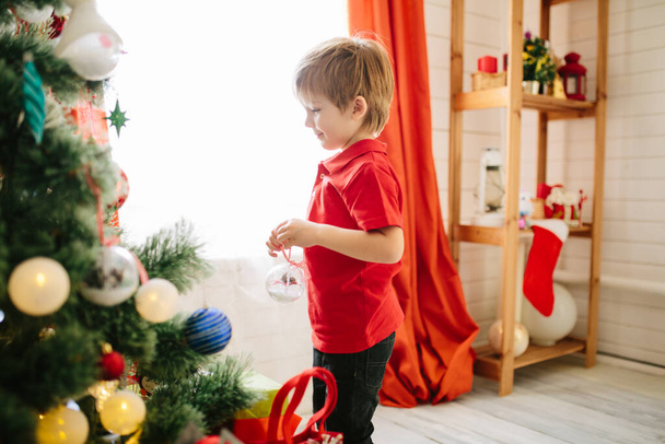 Cute little boy of about five year with blue eyes decorating a Christmas tree. - Foto, Imagen