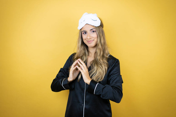 Young pretty woman wearing sleep mask and pajamas over isolated yellow background with Hands together and fingers crossed smiling relaxed and cheerful. Success and optimistic - Photo, Image
