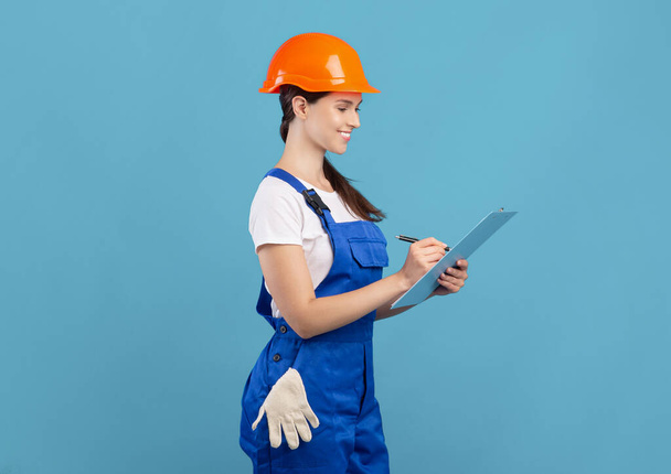 Usmívající se mladá údržbářka v kombinézách a na klobouku sestavující seznam stavebních materiálů - Fotografie, Obrázek