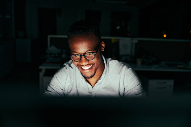 Успешный улыбающийся молодой африканский бизнесмен, работающий сверхурочно один за своим столом в офисе поздно ночью - Фото, изображение