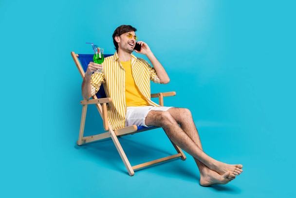 Dude i paradise. Full length photo man relax rest resort sit chaise-lounge drink cocktail call friend tell weekend wear white yellow shirt shorts barefoot isolated blue color background - Foto, Imagen