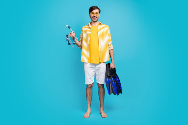 Coral reef i go. Full size photo positive guy traveler hold scuba diving gear flippers mask goggles tube wear yellow striped shirt white shorts barefoot isolated blue color background - Foto, Imagem