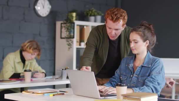 PAN de jeunes hommes d'affaires aux cheveux roux debout par bureau de collègues féminines travaillant sur un ordinateur portable et discutant du travail - Séquence, vidéo