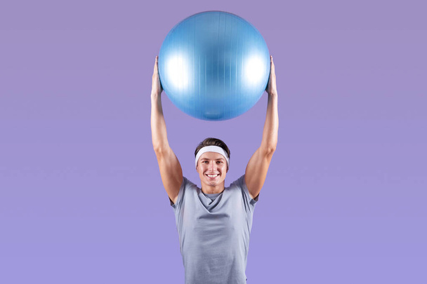 Radosny młody facet podnosząc fitball nad głową na fioletowym tle studio - Zdjęcie, obraz