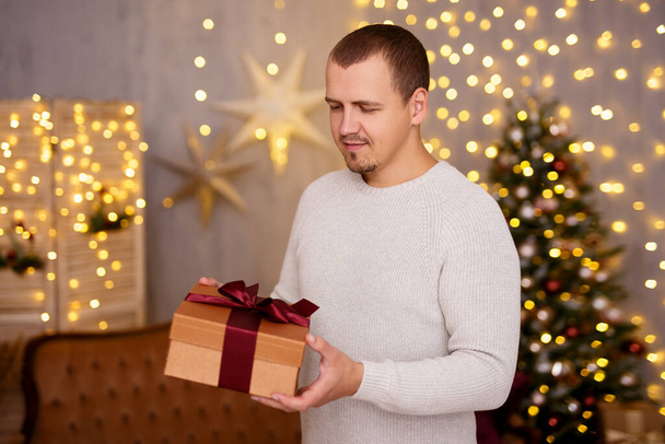 Weihnachts- und Neujahrskonzept - Porträt eines gutaussehenden Mannes mit Geschenkbox im geschmückten Wohnzimmer mit Weihnachtsbaum - Foto, Bild