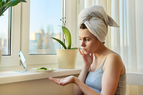 Young woman near home window looking in mirror, applying face cream - Photo, Image