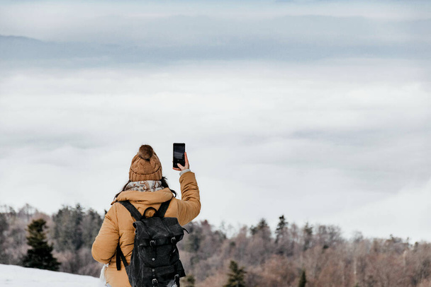 A young woman wearing warm clothes standing on the mountain and taking photos of a winter forest - Photo, Image