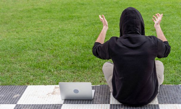 A young man wearing a hoodie sitting in a park with his laptop - Fotó, kép