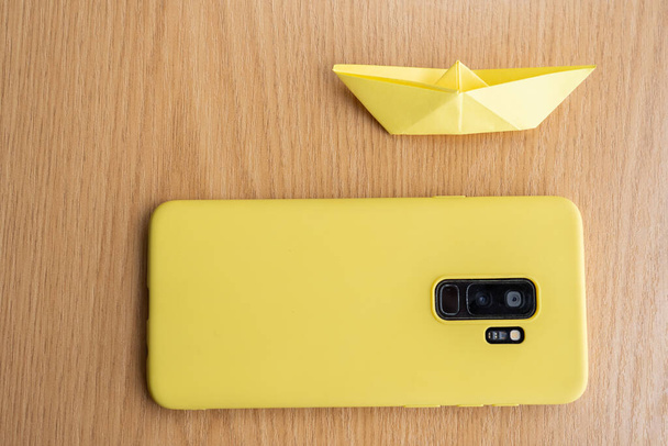 A high angle shot of a yellow cellphone and a paper boat on a wooden surface - Photo, Image