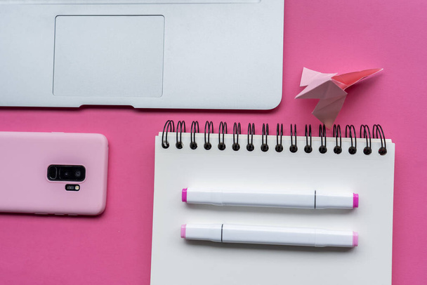 A  view of markers, notebook, laptop, phone, and swan origami on pink background - Foto, Imagen