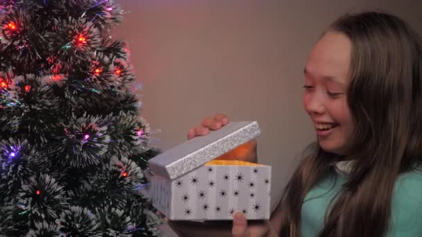 Una niña abre regalo de Santa Claus al lado del árbol de Navidad. Niño abre caja de regalo a la luz de guirnalda, se regocija y sonríe. Vacaciones y celebración de los niños de la familia, descanso de invierno. Feliz Navidad.. - Imágenes, Vídeo