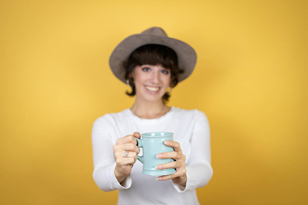 Junge kaukasische Frau mit Hut vor isoliertem gelben Hintergrund genießt und trinkt eine Tasse Kaffee und zeigt die Tasse in die Kamera - Foto, Bild