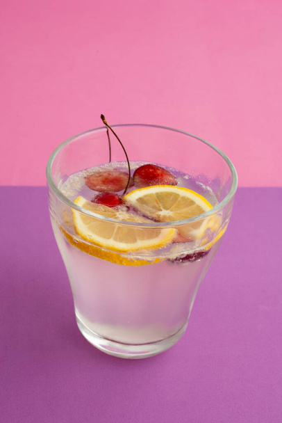 a glass of sparkling water cocktail with sliced lemon, cherry and berry. non-alcoholic drink, beverage. purple textured backdrop and pink background. carbonated water and bubble. abstract, close up. - Photo, Image