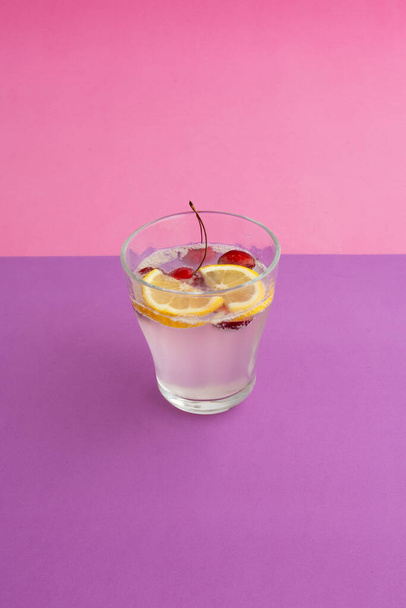 a glass of sparkling water cocktail with sliced lemon, cherry and berry. non-alcoholic drink, beverage. purple textured backdrop and pink background. carbonated water and bubble. abstract, close up. - Фото, изображение