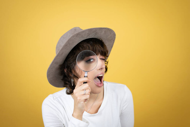Mujer joven caucásica con sombrero sobre fondo amarillo aislado sorprendida mirando a través de una lupa - Foto, imagen