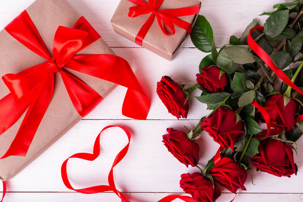 Concept de Saint-Valentin. Ruban rouge en forme de coeur, bouquet de roses et coffrets cadeaux enveloppés dans un pappeur kraft sur une table en bois blanc. - Photo, image
