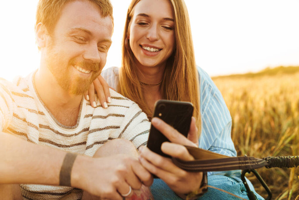 Image of a young happy cheery loving couple using mobile phone outside at the field while holding lead - Photo, Image
