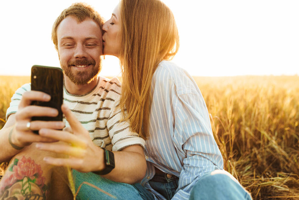 Image of a young positive loving couple taking a selfie by mobile phone outside at the field - Foto, Imagem