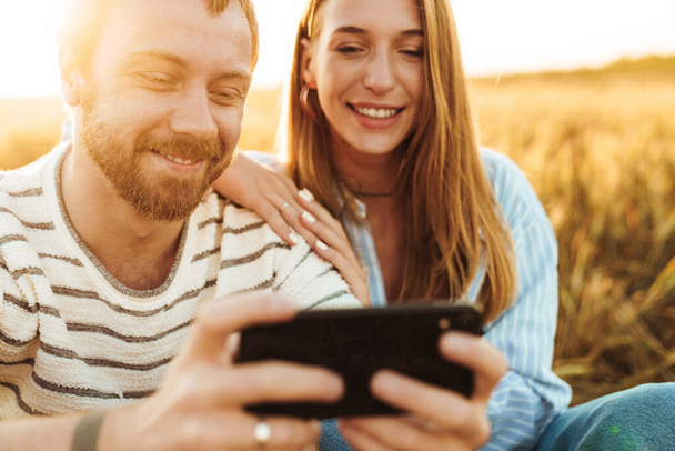 Image of a young happy positive loving couple using mobile phone outside at the field - Photo, Image