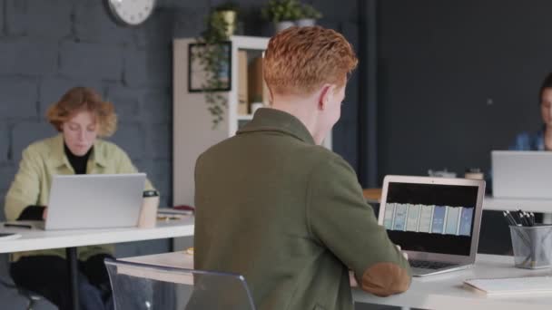 Vista posteriore PAN di giovane designer UI maschile seduto alla scrivania in ufficio e guardando nuovi modelli di interfaccia app - Filmati, video