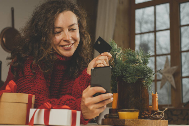 Close up happy brunette woman holding bank credit card and looking at smartphone, making purchase before New Year, sitting at table at home. Paying online for Christmas gifts, enjoying winter holiday - Foto, Imagen