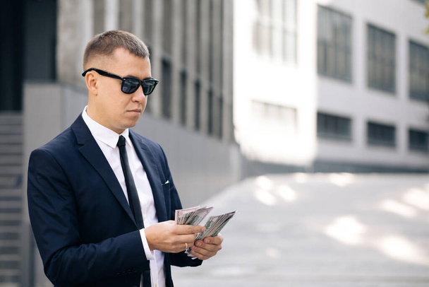 Portrait of handsome caucasian young businessman in sunglasses counting money while standing outdoors. Joyful male with many dollars. Luxury life. Money concept - Foto, afbeelding