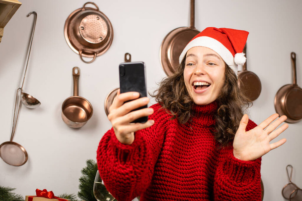 Felice donna caucasica sorridente in cappello rosso di Natale e maglione con il telefono, guardando lo schermo, agitando la mano e salutando a casa cucina. Chiacchierare online con amici o parenti, fare videochiamate - Foto, immagini