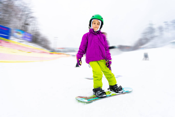 Snowboard Winter Sport. Το κοριτσάκι μαθαίνει σνόουμπορντ, φορώντας ζεστά χειμωνιάτικα ρούχα. Χειμερινό υπόβαθρο. - Φωτογραφία, εικόνα