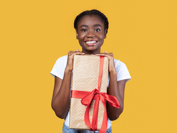 Fröhliche schwarze Dame mit Geschenkbox posiert vor gelbem Hintergrund - Foto, Bild