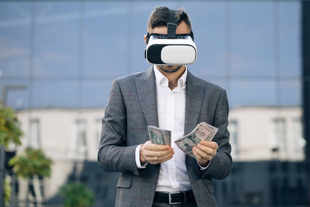 Rich man wearing virtual reality headset counting money. Young businessman standing with pack of dollars near modern office building. Richness and success concept. 3D VR Glasses - Foto, Imagen