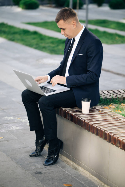 Distance working. Isolated man in a suit. Handsome young businessman sitting outdoors working with his laptop. Crisis 2020. Coronavirus outbreak - Photo, Image