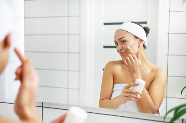 A young smiling woman wrapped in towels holding moisturizing cream and applying it on her face in bathroom. - Photo, Image