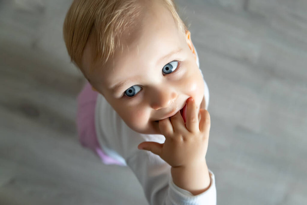 Cute adorable infant blond happy little toddler boy stand in home room looking above and sucking hand in mouth against floor background. Child virus and sick runny nose. Kid healthcare concept. - Photo, Image
