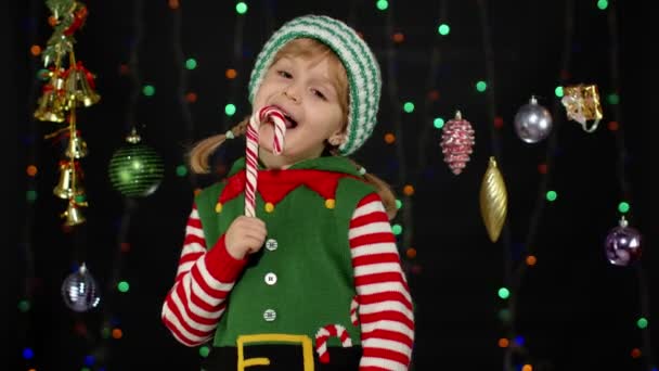 Kid girl w Boże Narodzenie elf Santa Claus pomocnik kostium lizanie cukierki trzciny cukrowej lizak karmel słodycze - Materiał filmowy, wideo
