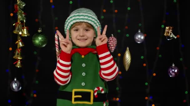 Blondýny dítě dívka v vánoční skřítek Santa Claus helper kostým dělat mír gesto, ukazuje v znamení - Záběry, video
