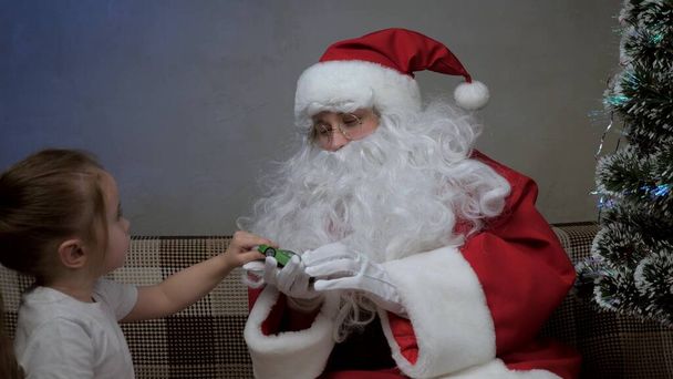Άγιος Βασίλης κάθεται στον καναπέ με ένα μικρό κορίτσι, Άγιος Βασίλης έδωσε ένα δώρο στο παιχνίδι αυτοκίνητο, παιδί χαίρεται και αγκαλιάζει μαγικό παππού. Γιορτή και γιορτή. Οικογενειακές χειμερινές διακοπές. Ευτυχισμένη παραμονή Χριστουγέννων. - Φωτογραφία, εικόνα