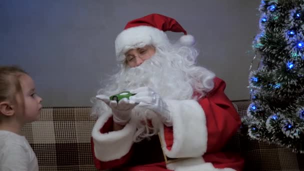 Santa Claus sits on couch with a little girl, Santa gave a gift to toy car, child rejoices and hugs magic grandfather. holiday and celebration. Family childrens winter vacation. Happy Christmas Eve. - Footage, Video