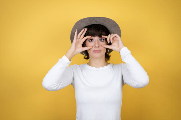 Jonge blanke vrouw draagt hoed over geïsoleerde gele achtergrond Proberen om ogen te openen met vingers, slaperig en moe voor de ochtend vermoeidheid - Foto, afbeelding