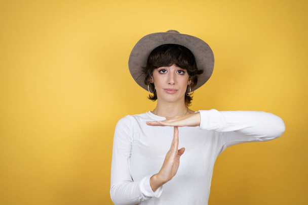 Fiatal kaukázusi nő visel kalap felett elszigetelt sárga háttér csinál idő out gesztus kezével, frusztrált és komoly arc - Fotó, kép