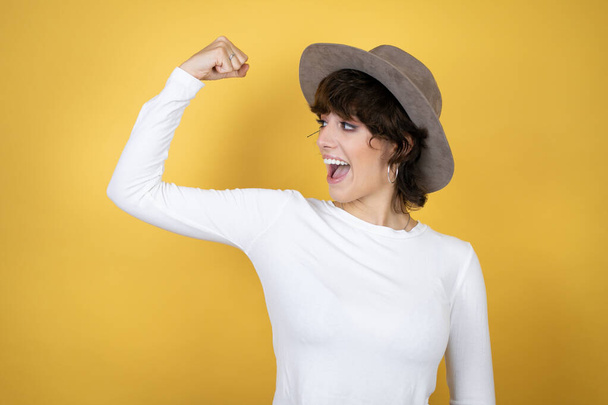 Jonge blanke vrouw draagt hoed over geïsoleerde gele achtergrond tonen armen spieren glimlachen trots - Foto, afbeelding